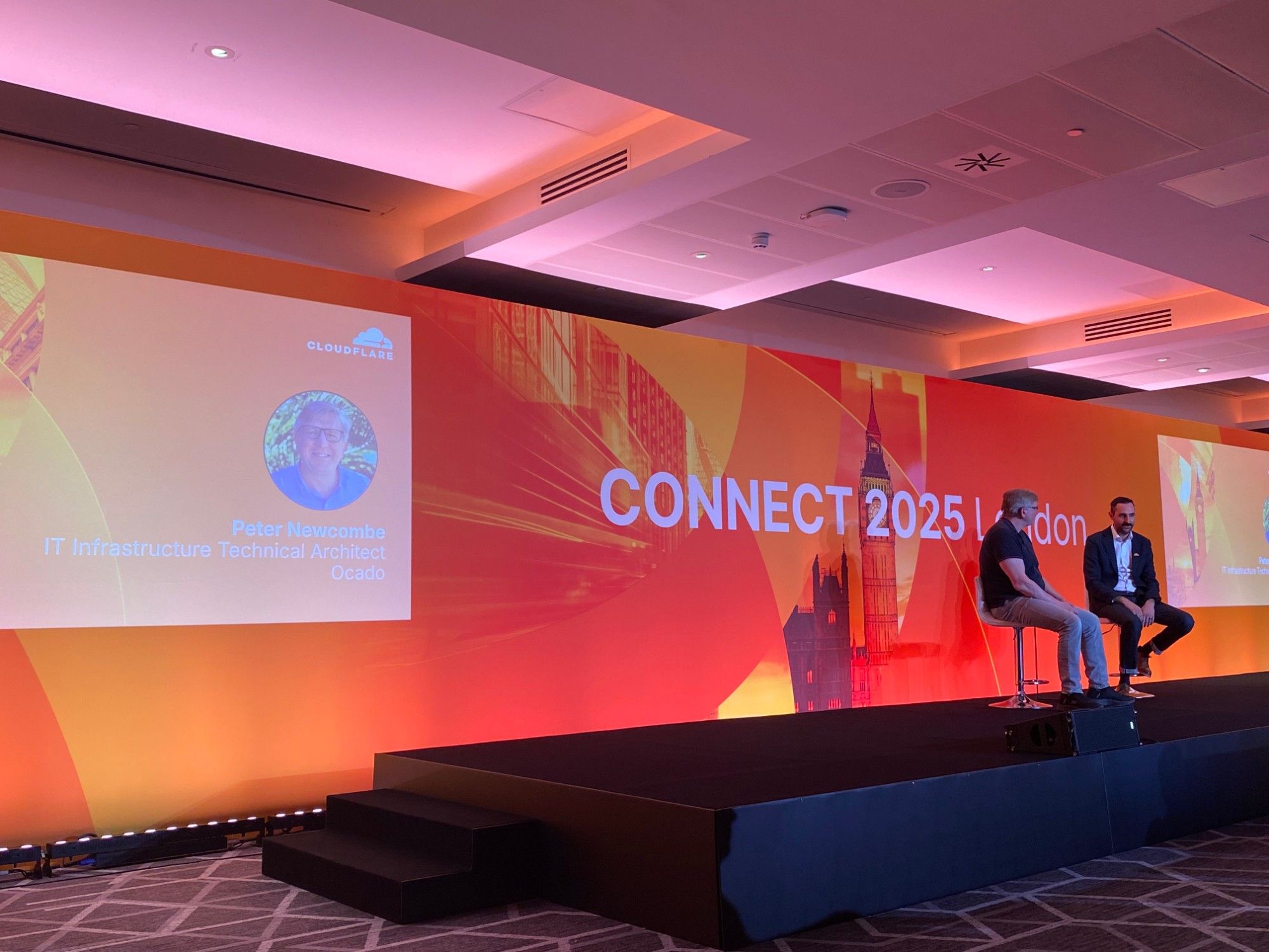 Pete Newcombe on stage in a two-person fireside chat at Cloudflare Connect London 2025, beside a large screen showing his name, the title IT Infrastructure Technical Architect, and Ocado.